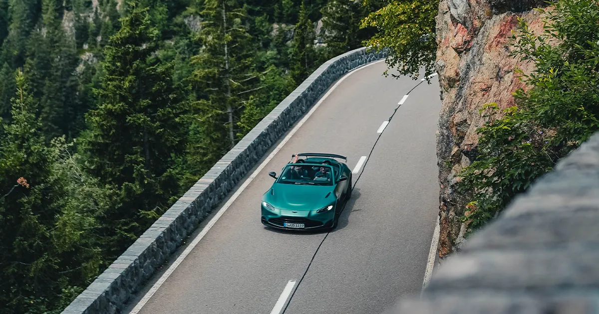 A teal Aston Martin Vantage F1 Roadster being driven with its top down by two HunterMoss guests.