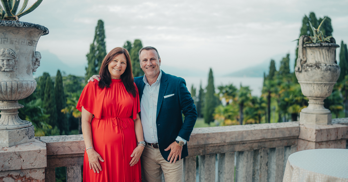 A happy couple in elegant outfits on a beautiful stone balcony with cypress trees in the grounds behind