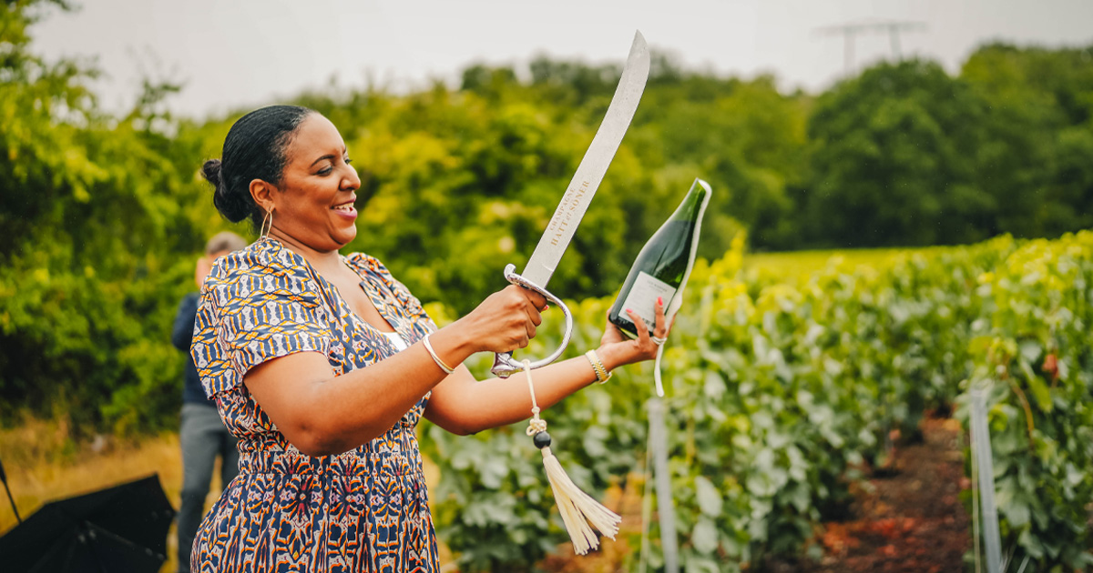 A lady smiles as she removes the cord from a champagne bottle using a traditional sabre
