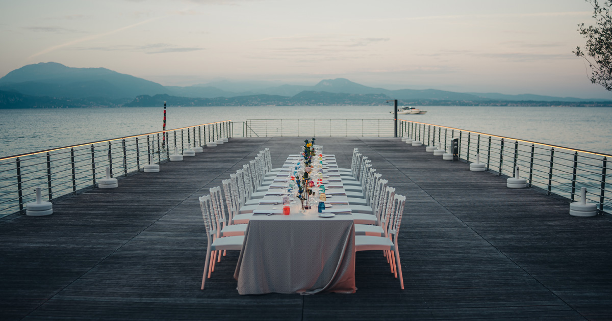 A long dining table is set on a jetty on a lake with mountains in the background