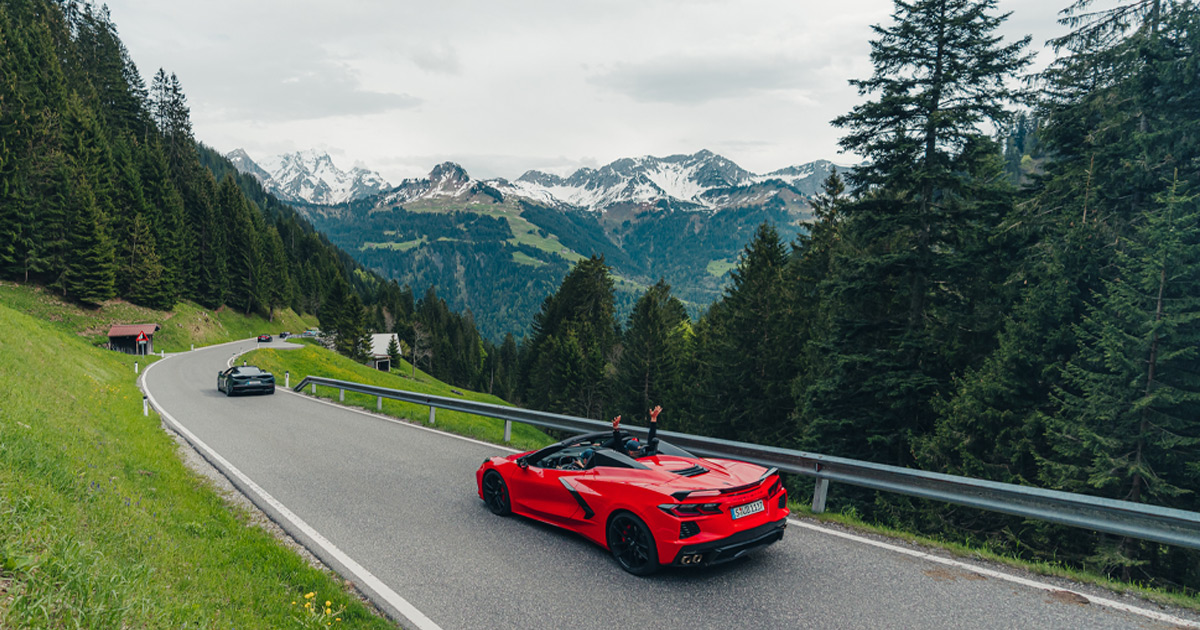 A red Corvette C8 convertible driving along an alpine road with mountains in the distance