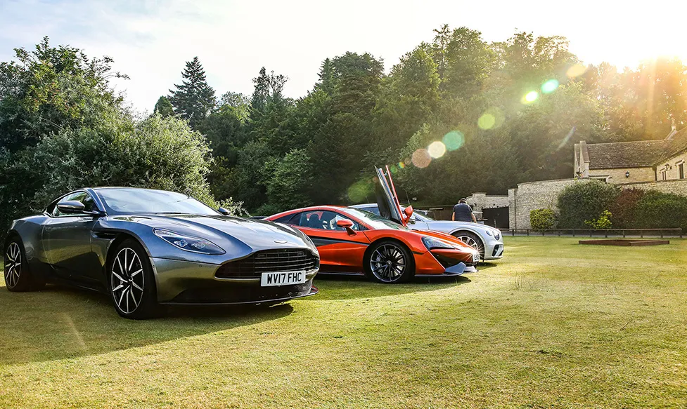 A brand new Aston Martin DBS belonging to the HunterMoss fleet sits next to a red McLaren on a grassy lawn in the afternoon sun