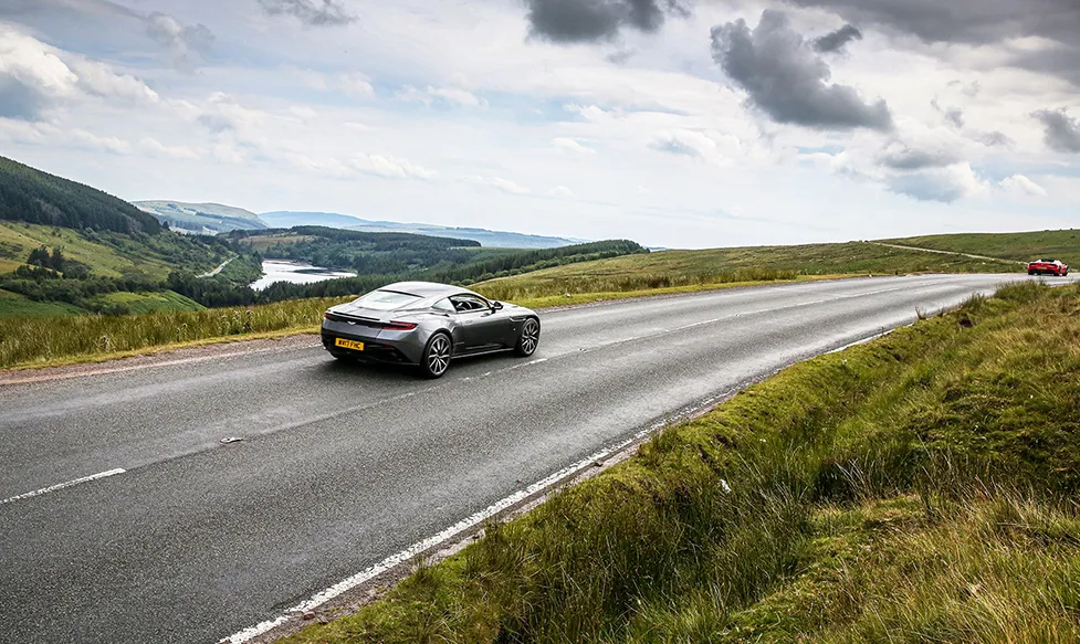 A gunmetal grey, late-model Aston Martin DBS crests a hill in the English countryside on an HunterMoss supercar holiday