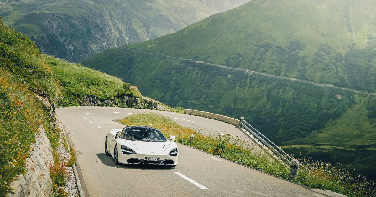 Join us to enjoy motoring at its very best. A white McLaren 720S exits a hairpin corner on an alpine pass during an HunterMoss experience.